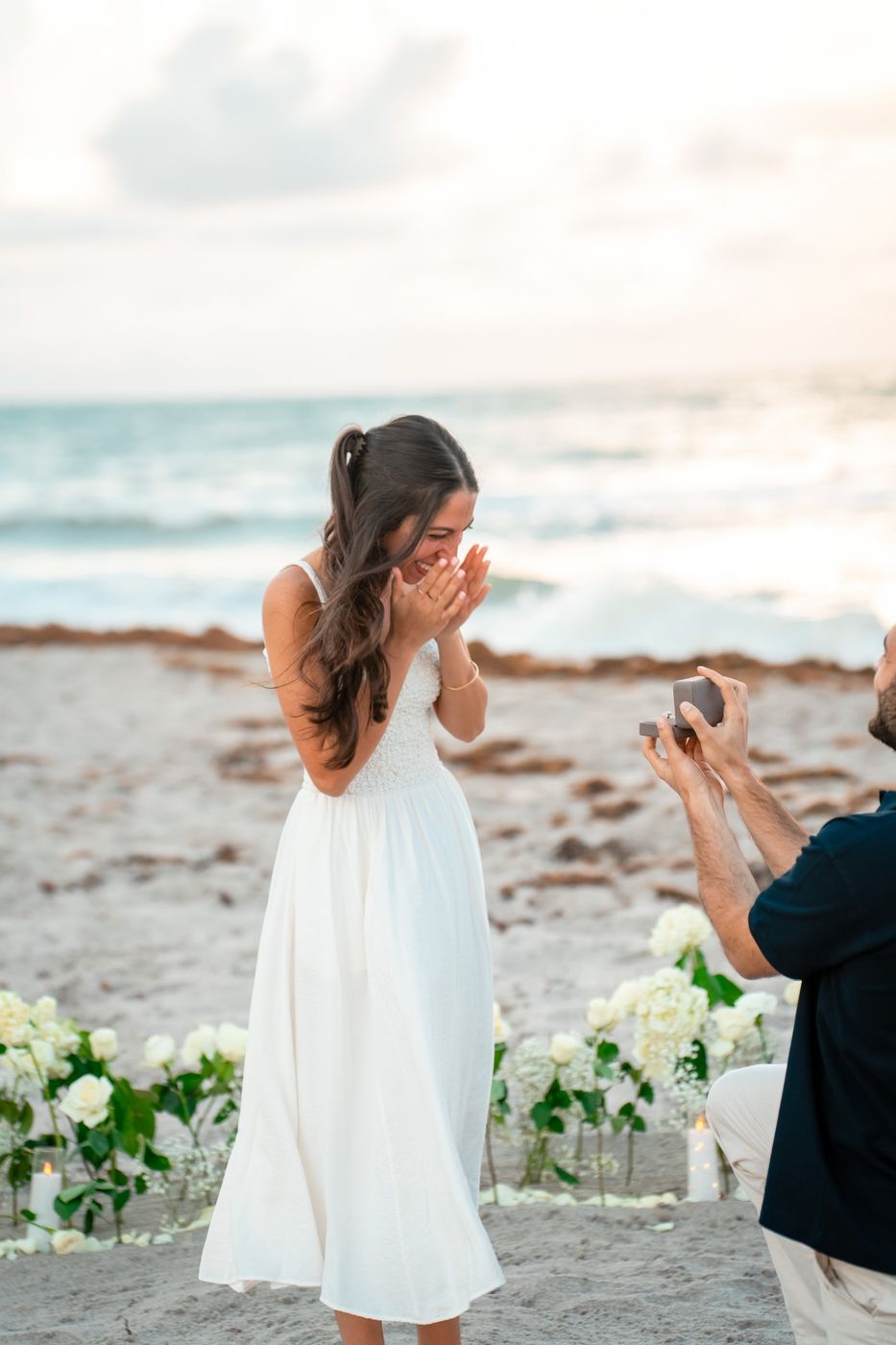 Talya laughing with her hands at her face as Sahar kneels with a ring on the beach